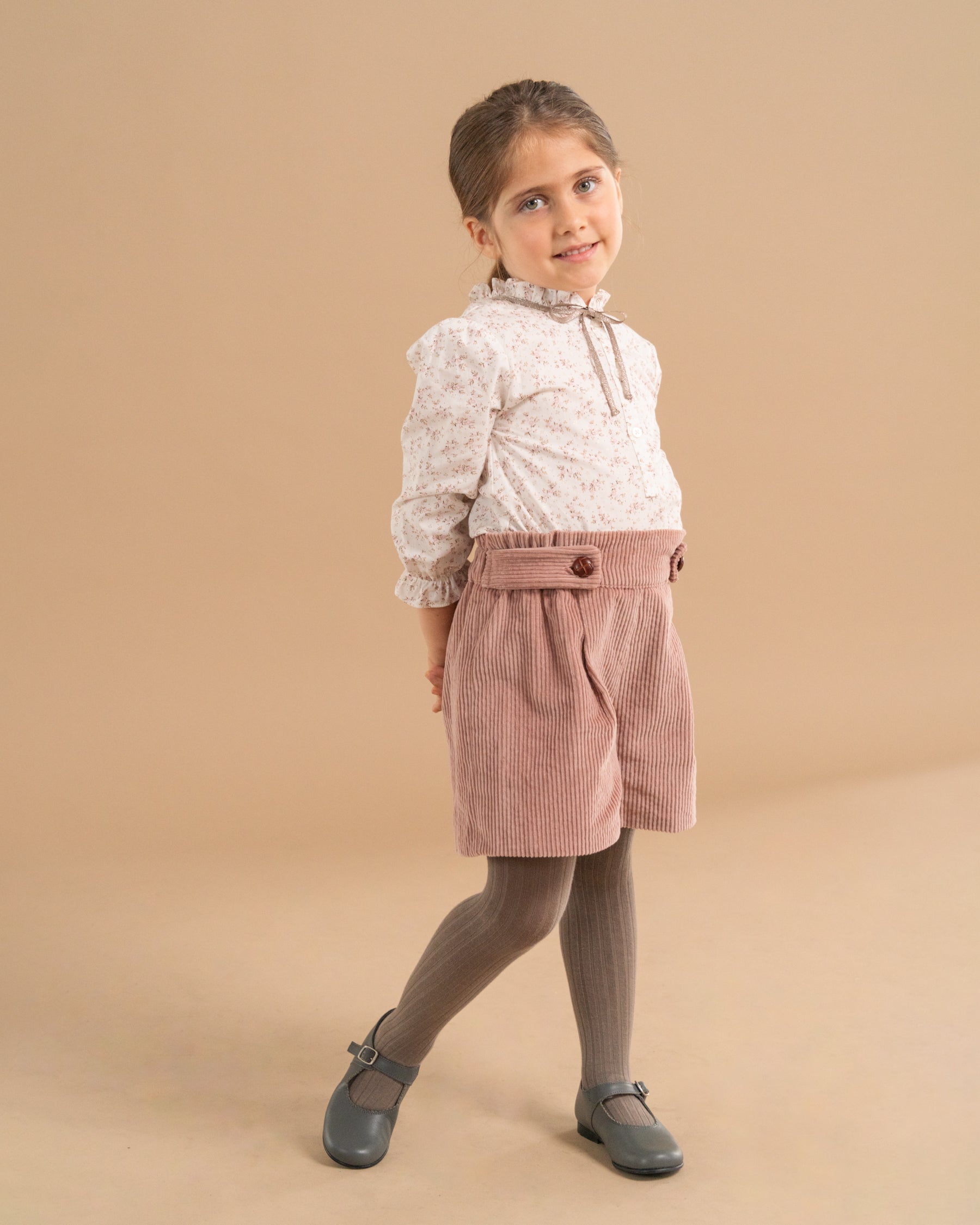 A young girl modeling a two-piece outfit consisting of a white floral long-sleeve blouse and dusty rose corduroy shorts with a matching fabric belt, paired with dark gray tights and gray Mary Jane shoes, set against a solid beige studio background.