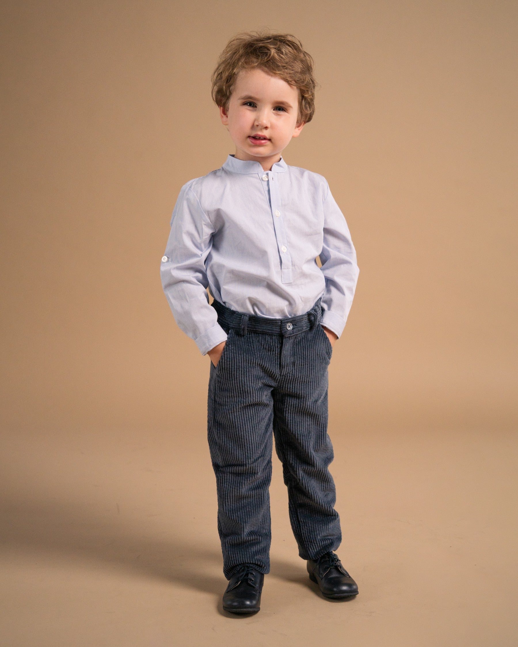A toddler boy modeling a light blue long-sleeve shirt with a mock neck and zipper placket, paired with dark blue pinstriped trousers and black shoes, against a solid tan studio background.