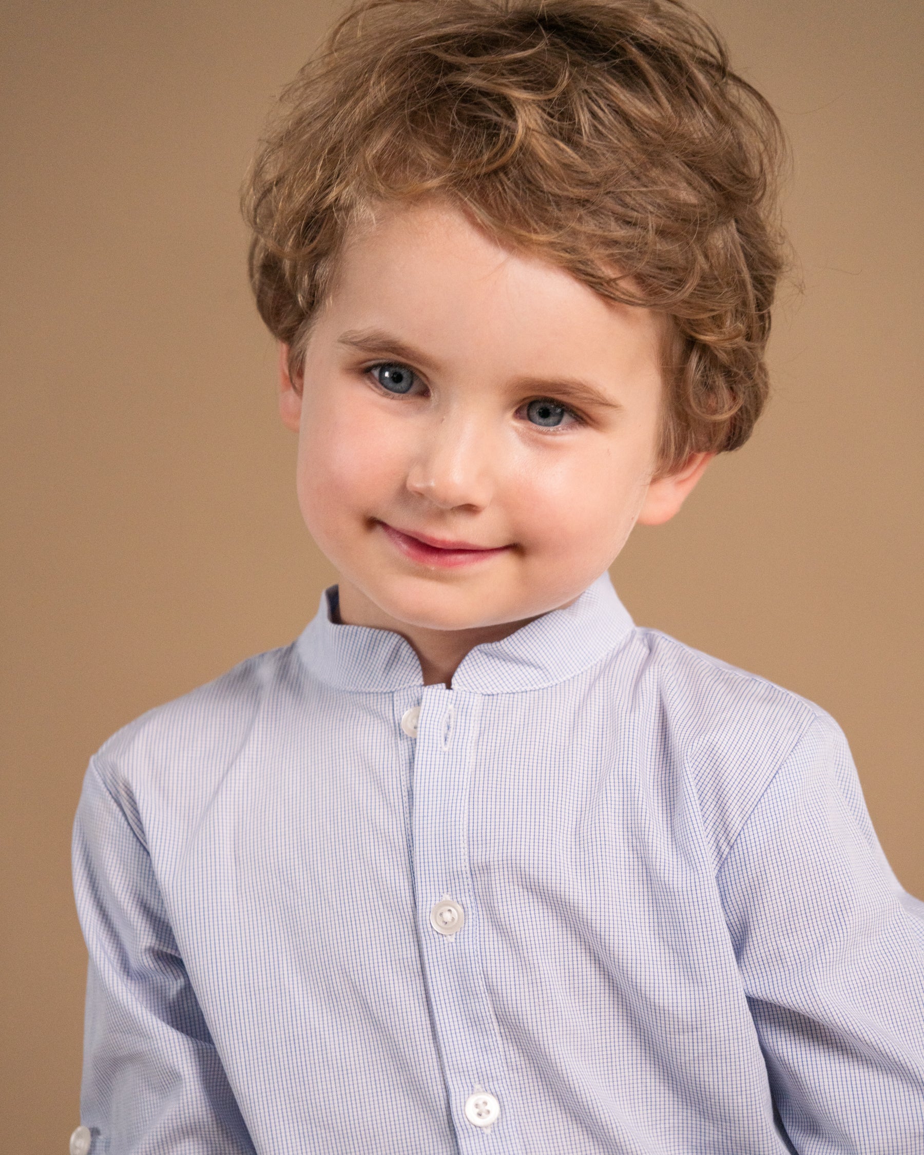 A young child with curly light brown hair wearing a light blue and white micro-striped button-up shirt, photographed against a solid warm beige background.
