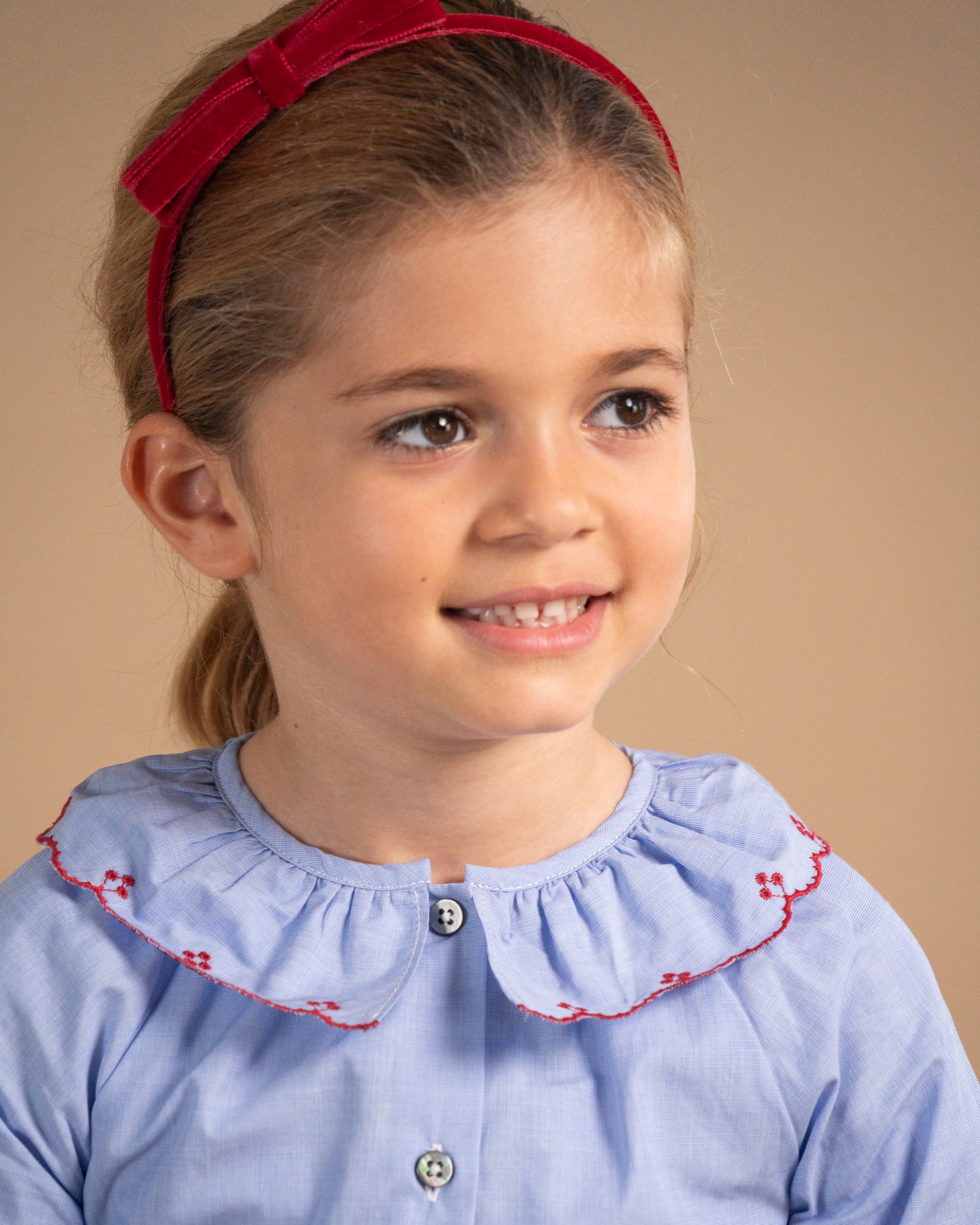 A young girl with light brown hair wearing a red velvet bow headband and a light blue button-up shirt with a ruffled Peter Pan collar trimmed in red embroidery, set against a solid beige background.