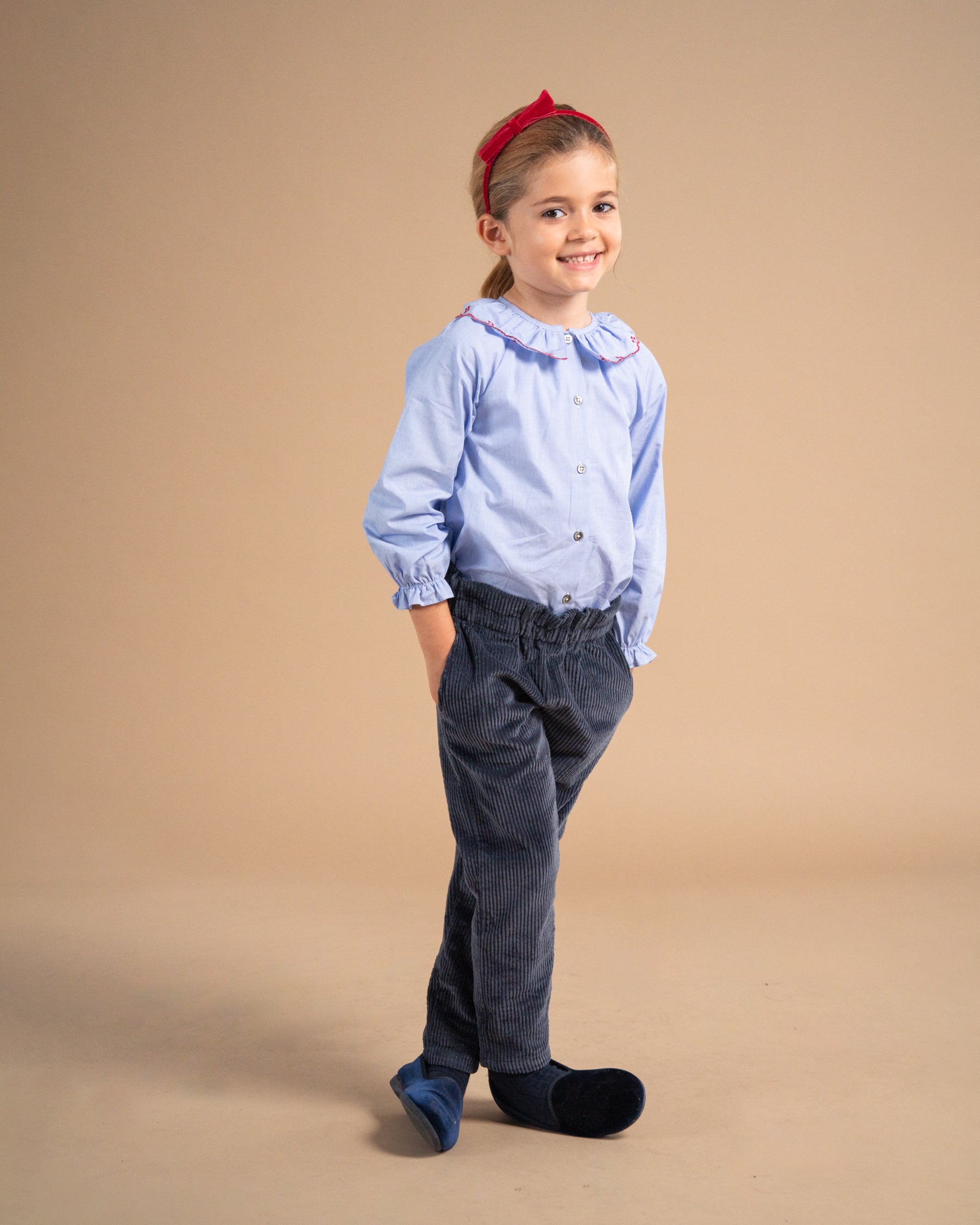 A young girl modeling a light blue long-sleeve button-up shirt with a ruffled collar, paired with dark corduroy trousers and a red bow headband, standing against a solid beige studio background.