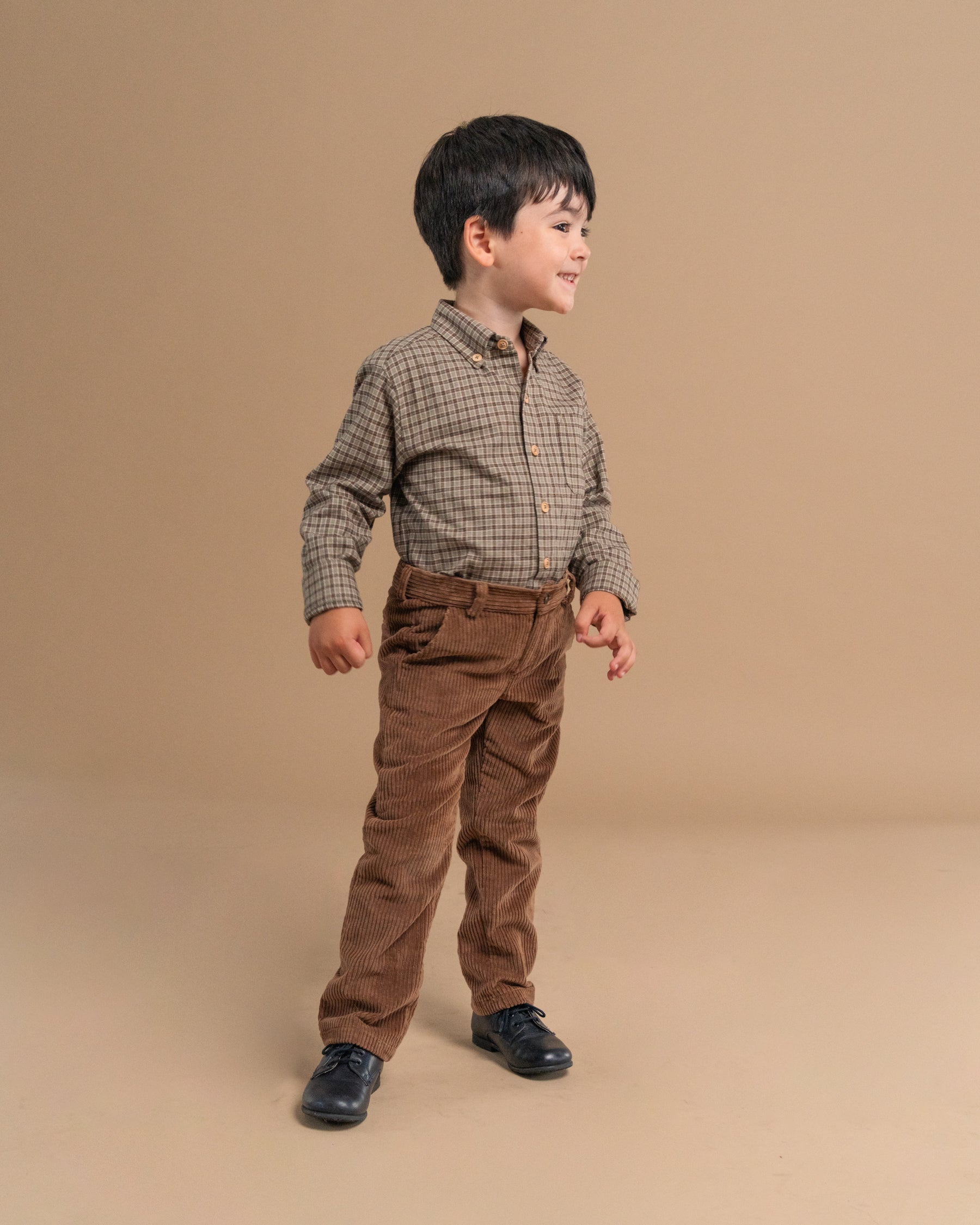 A young boy modeling a long-sleeve button-down shirt in a brown and beige checkered pattern, paired with brown corduroy pants and black shoes, standing against a solid beige studio backdrop.