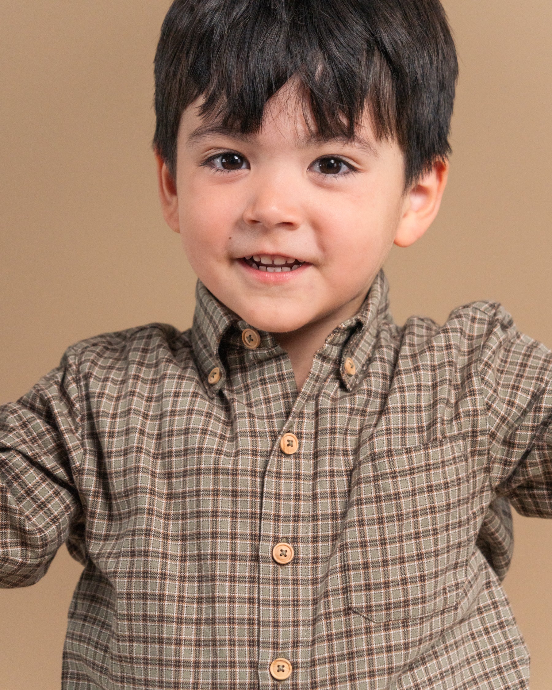 A young boy smiling, wearing a brown and beige plaid button-down shirt, photographed against a solid tan studio background.