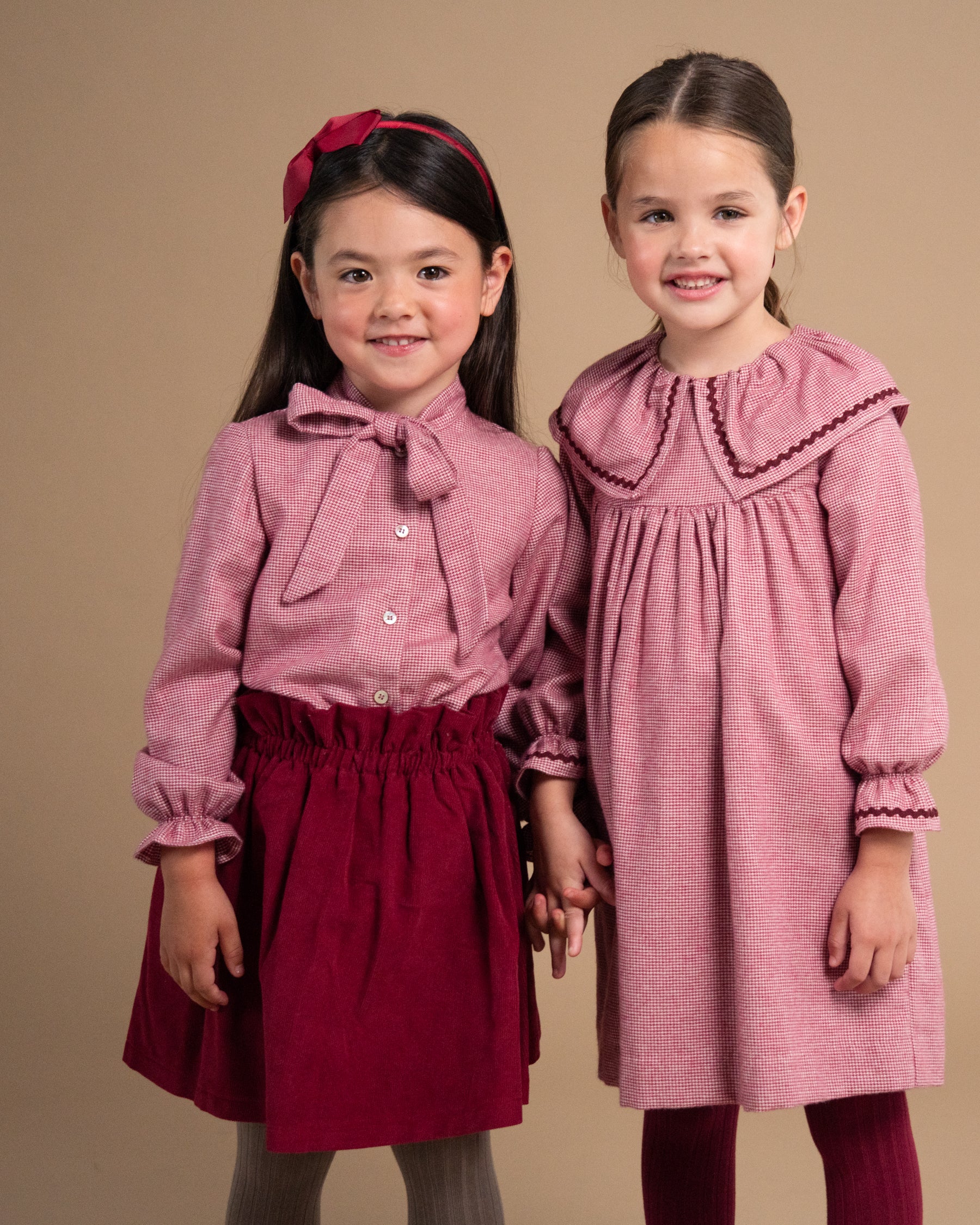 Two young girls modeling children's dresses against a solid beige studio background. The girl on the left wears a dusty rose long-sleeve blouse with a bow collar and a dark red skirt, holding the hand of the girl on the right who wears a matching dusty rose dress with a large Peter Pan collar and dark red trim.
