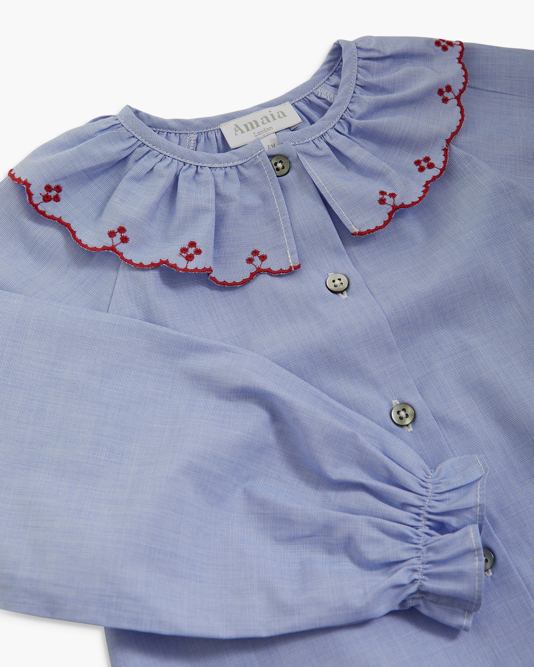 A close-up of a light blue long-sleeve blouse with a ruffled collar trimmed in red embroidery and decorative buttons.