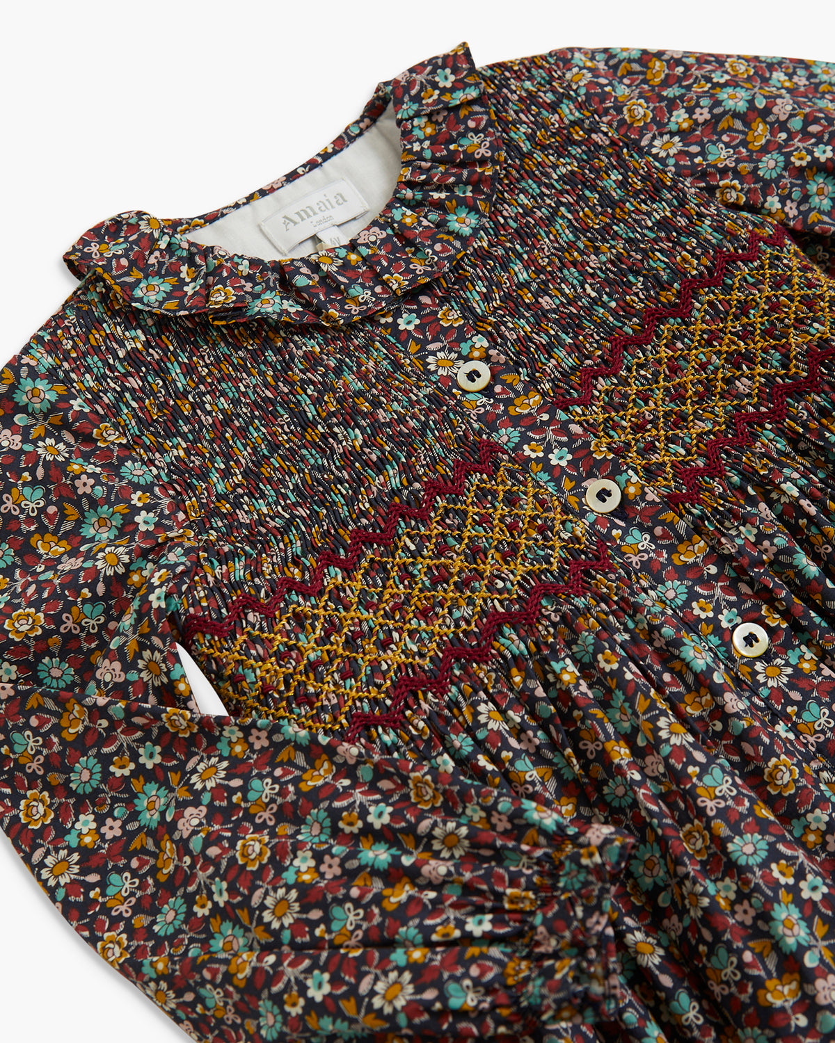 A close-up of a long-sleeve blouse with a smocked bodice, featuring a dense multicolor floral print in purple, yellow, blue, and white, accented by a burgundy and yellow embroidered chevron pattern across the chest. The brand name 'Amigo' is visible on the inner neck label.