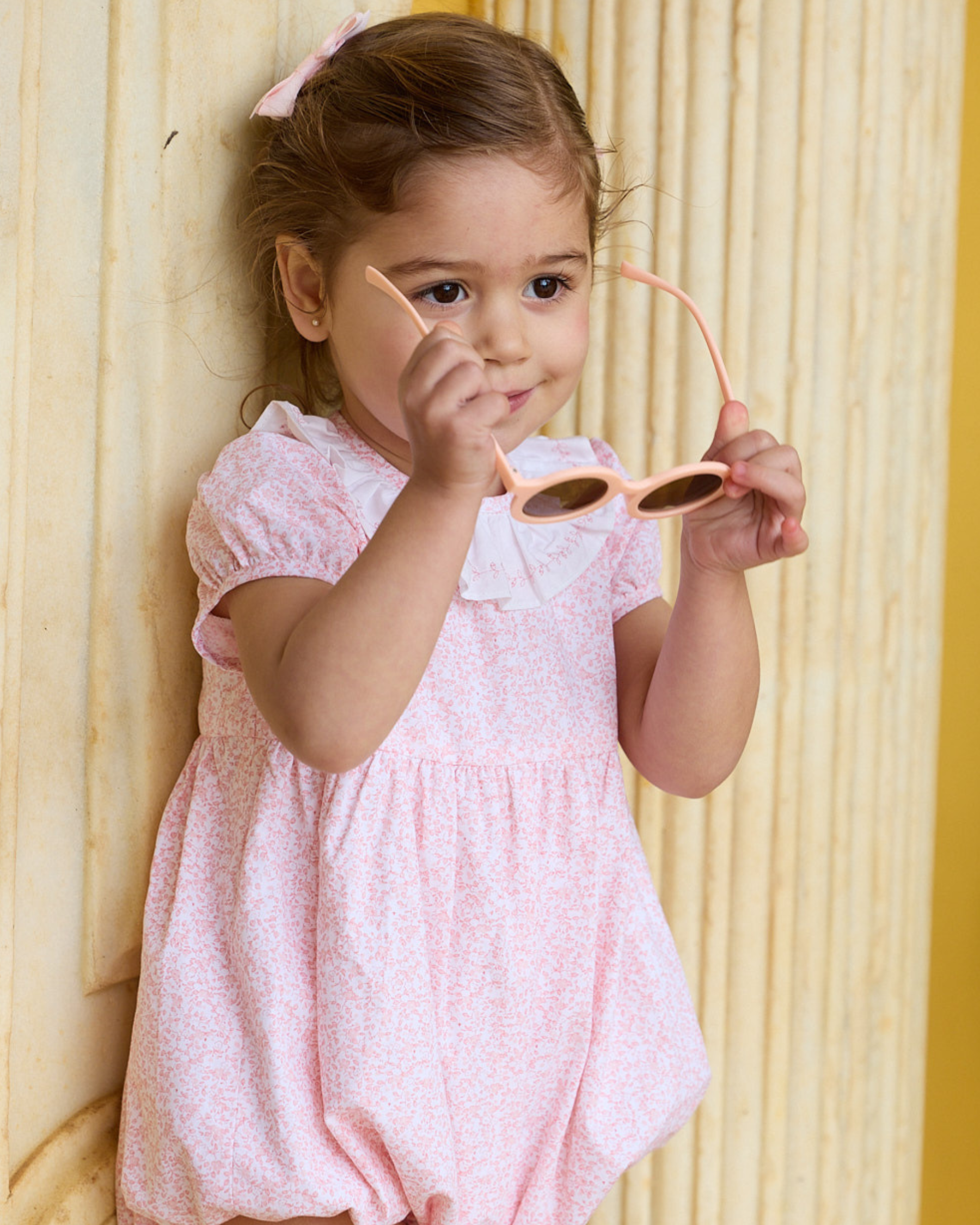 Young girl in a pink dress holding peach-colored sunglasses against a yellow background