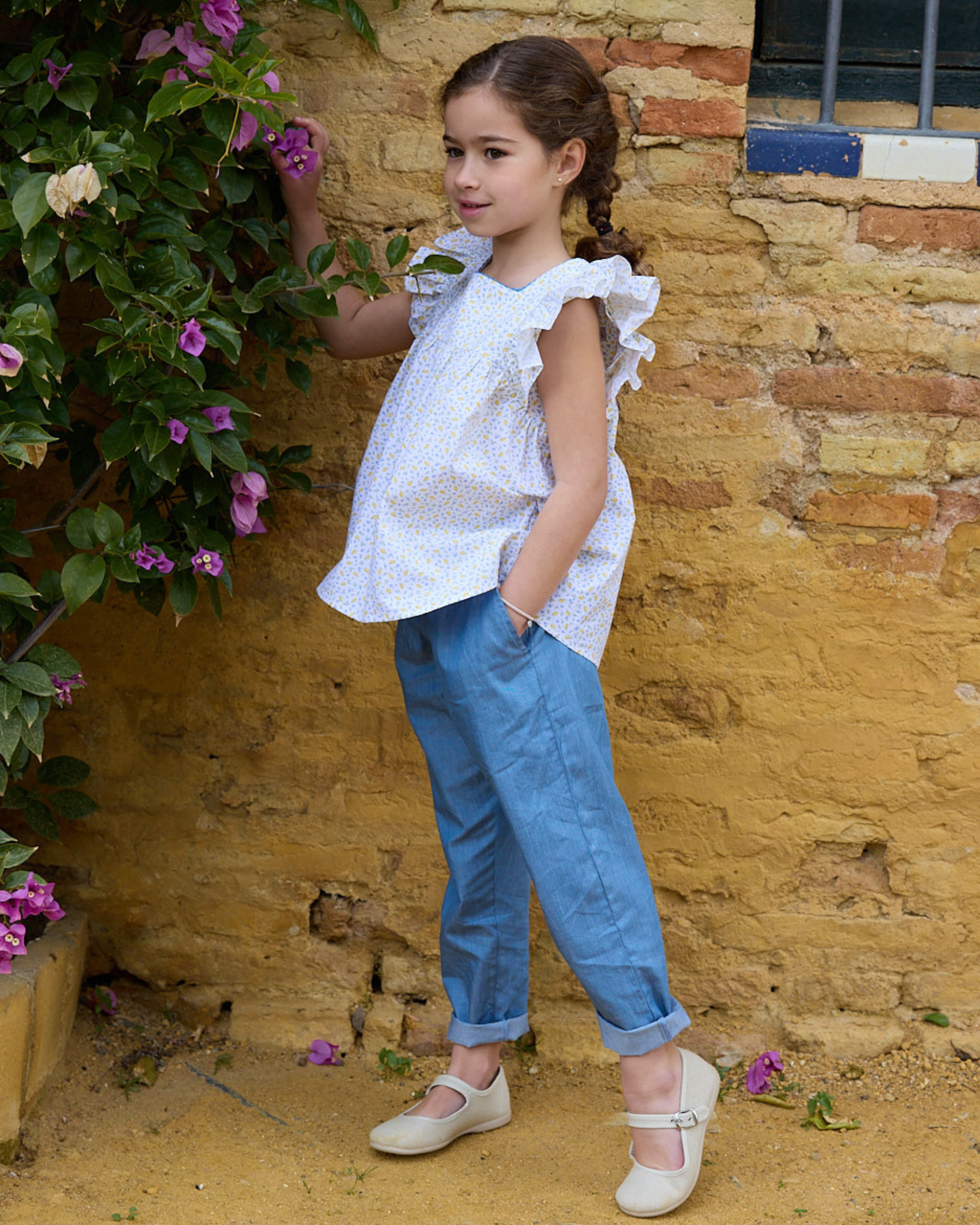 Young girl in a white blouse and blue pants standing against a rustic brick wall with flowers.