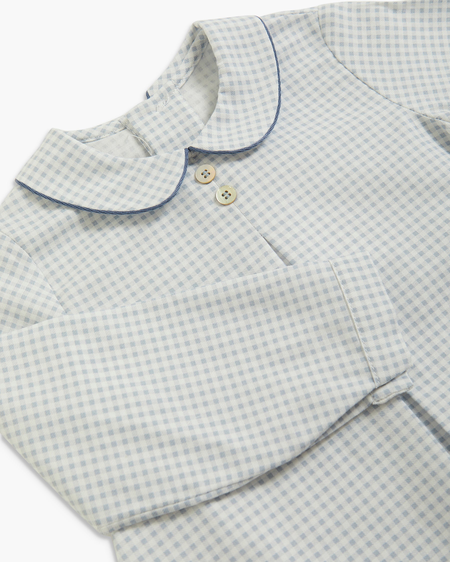 A close-up of a children's long-sleeve top with a light blue and white gingham pattern, featuring a Peter Pan collar with dark blue piping and two cream buttons.