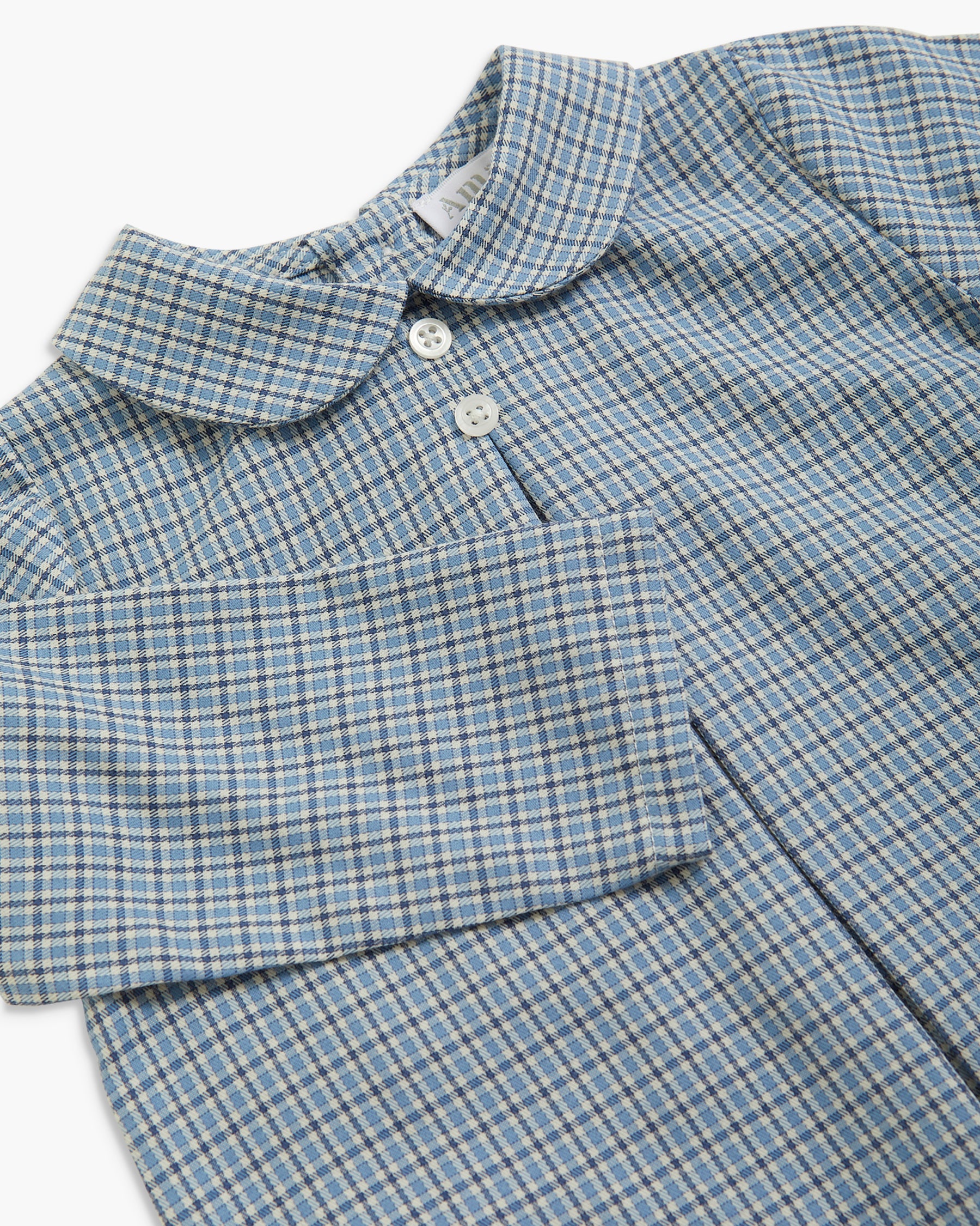 A close-up of a long-sleeve children's shirt with a blue, gray, and white micro-check pattern, featuring a Peter Pan collar and white buttons.
