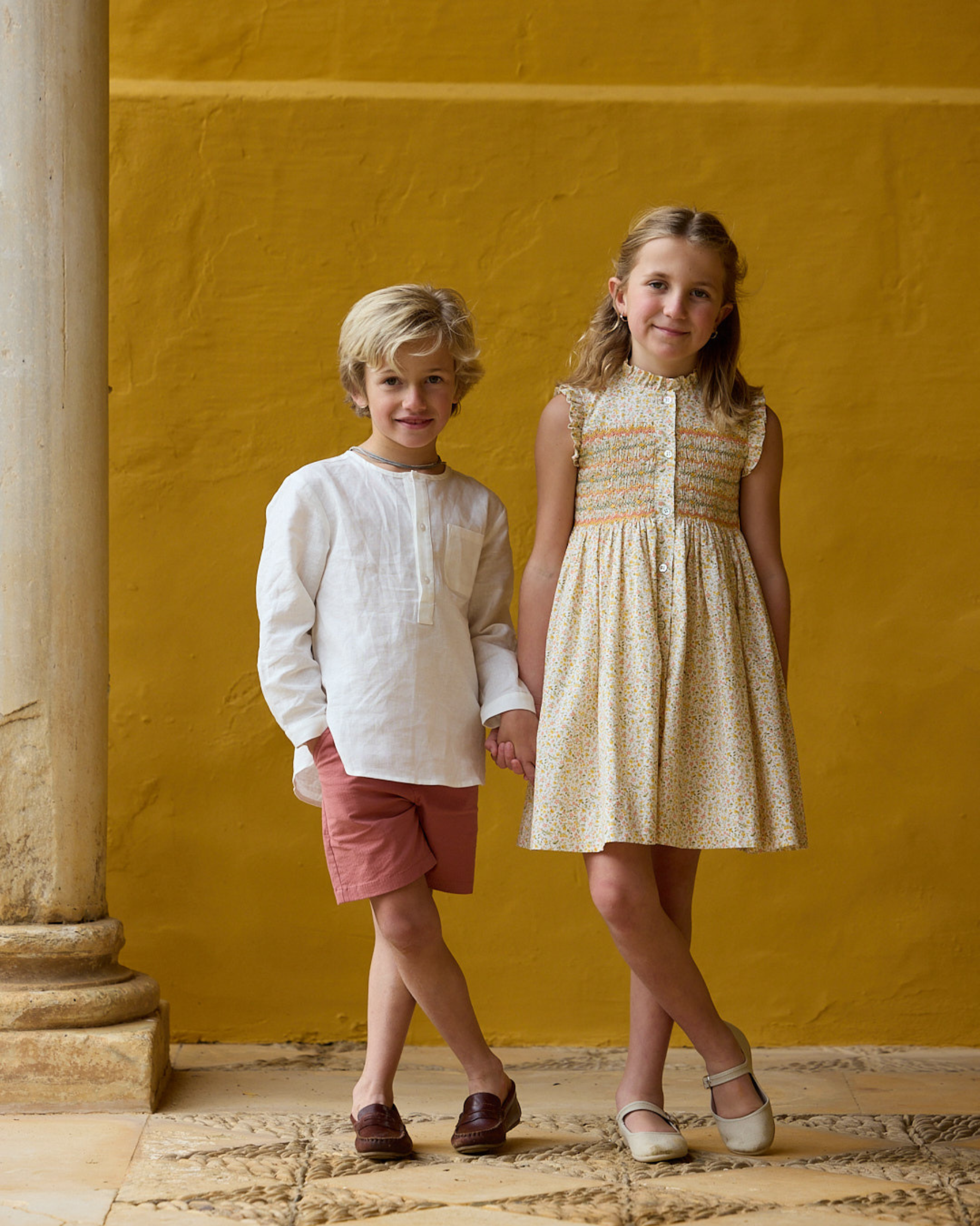 Two children standing against a yellow wall with a stone column.