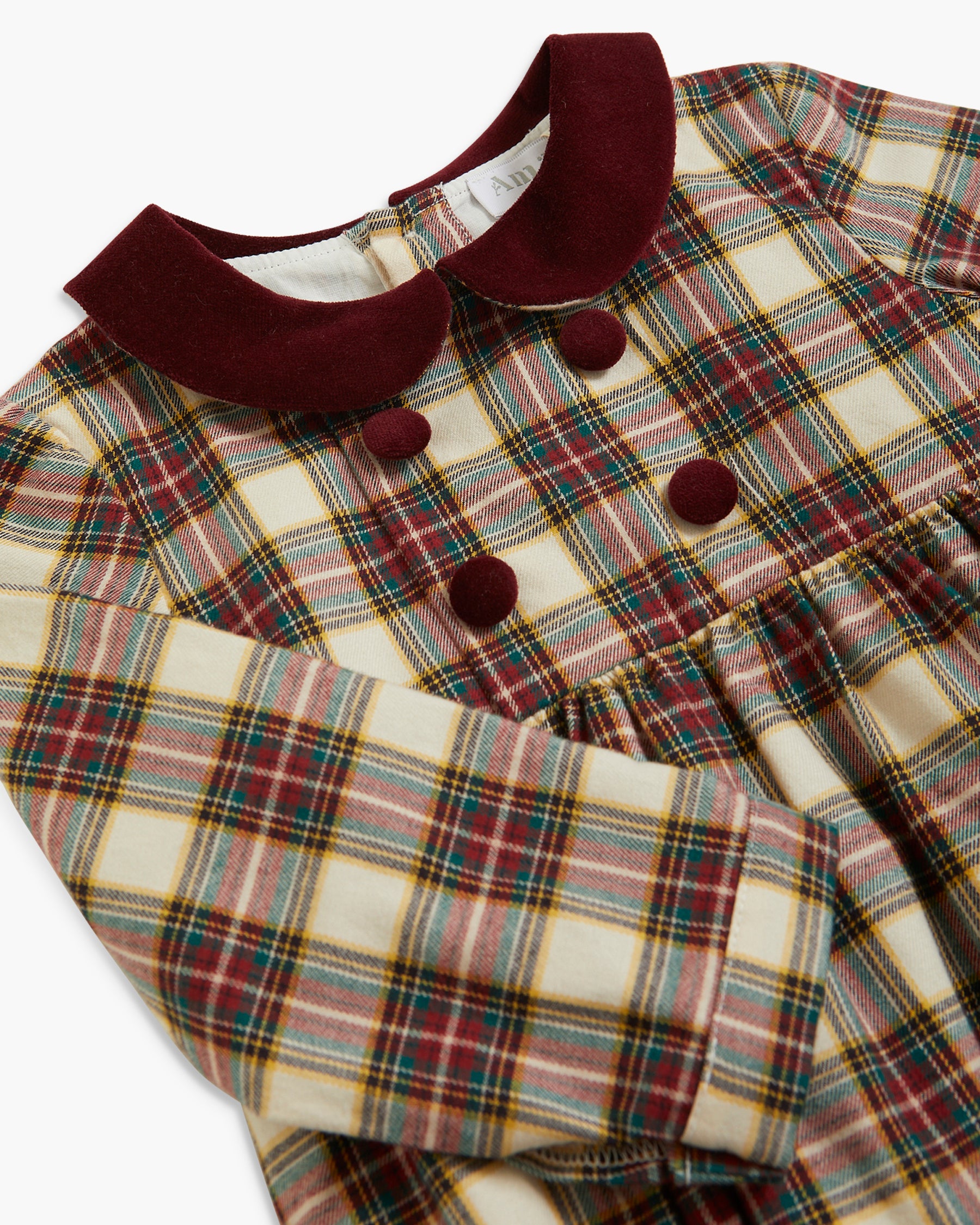 A close-up of a long-sleeve plaid dress featuring a cream base with red, green, and yellow stripes, accented by a contrasting dark burgundy velvet Peter Pan collar and matching buttons.