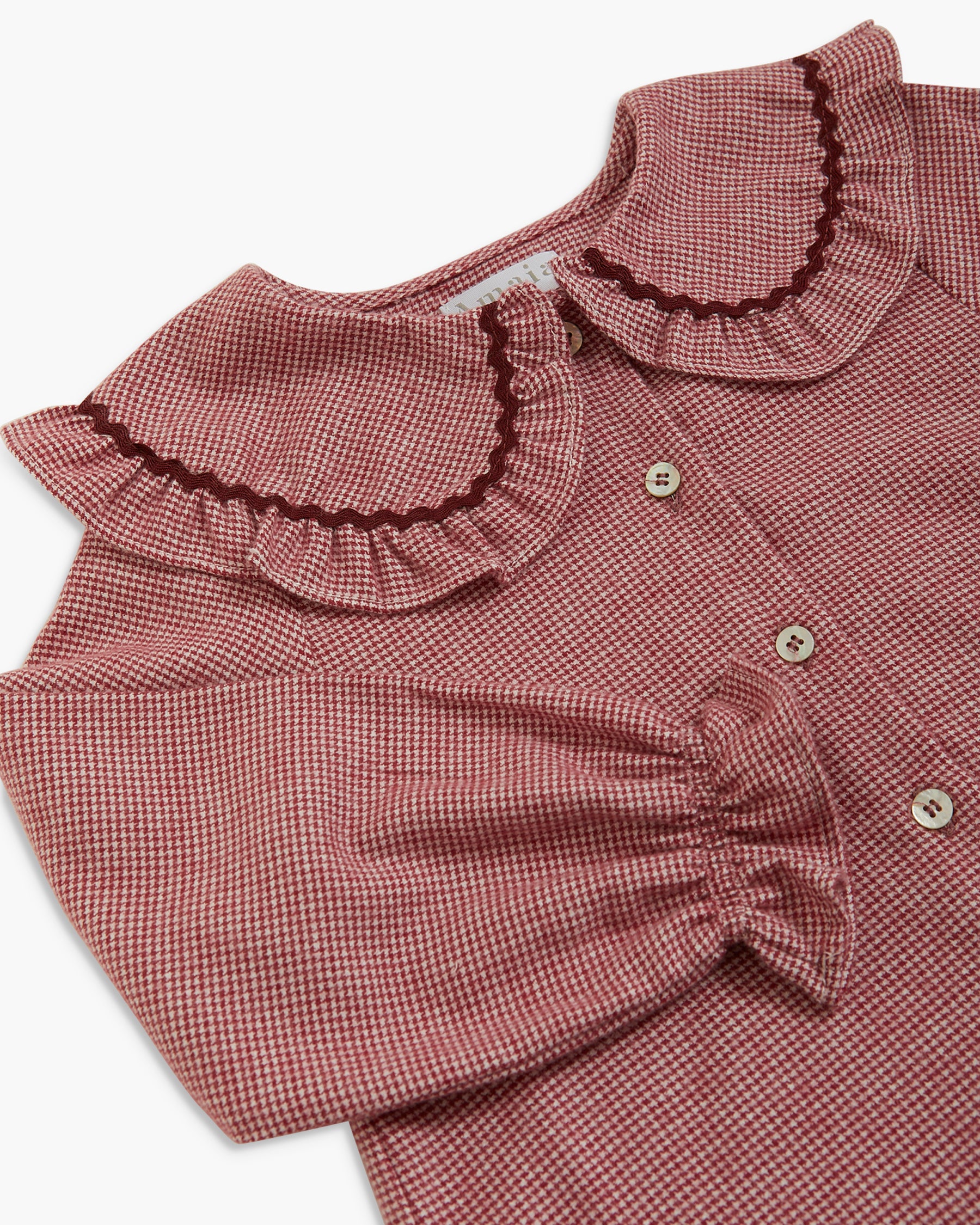 A close-up of a children's blouse with a dusty rose and white micro-check pattern, featuring a large ruffled Peter Pan collar with dark trim, buttoned front, and a gathered sleeve detail.