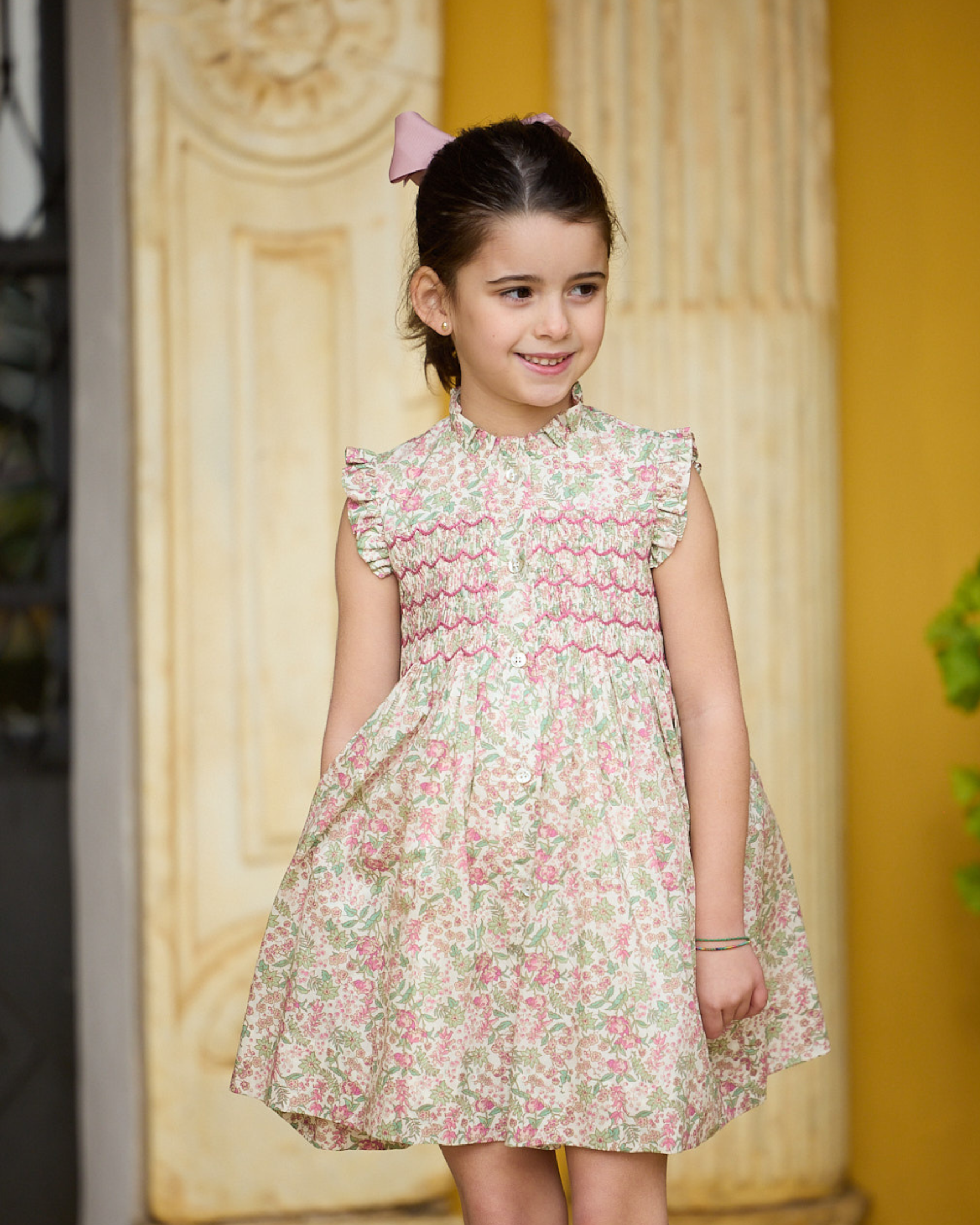 Young girl wearing a floral dress standing against a decorative wall.