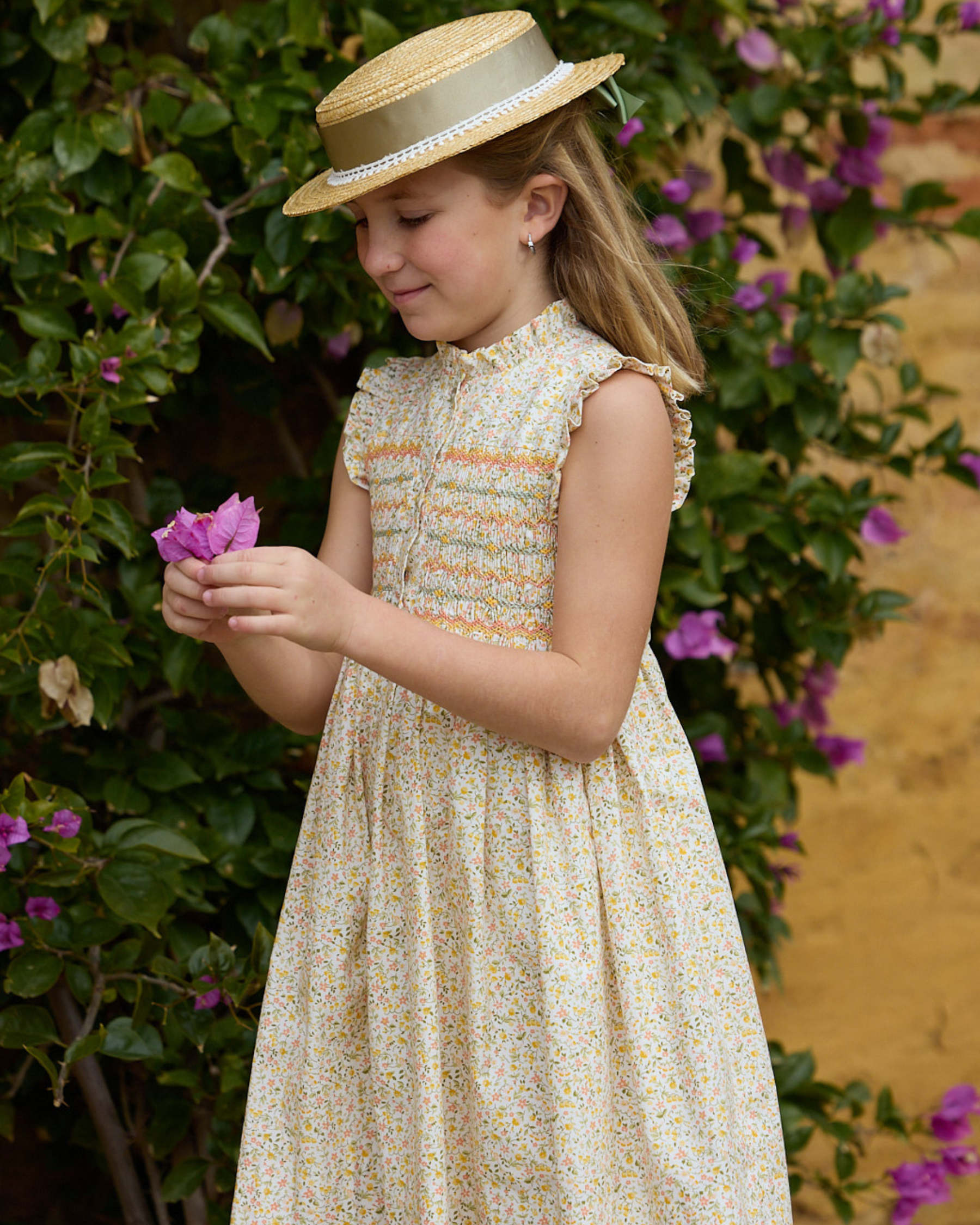 Young girl in a floral dress and straw hat holding a flower, standing against a floral background.
