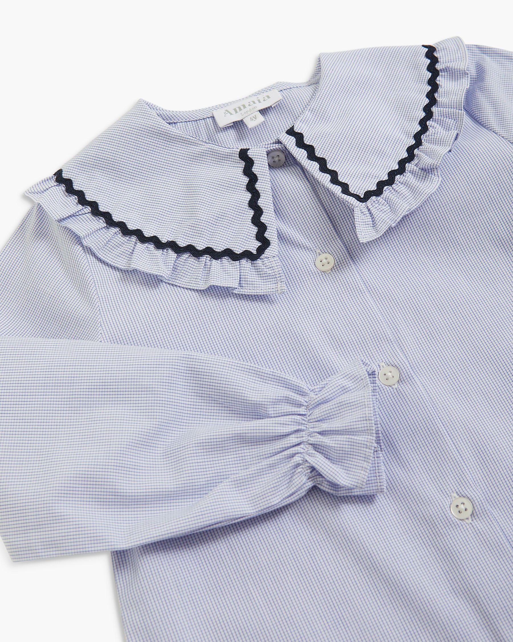 A close-up of a children's long-sleeve blouse with a light blue and white micro-striped pattern, featuring a large ruffled collar trimmed with black scalloped edging and a gathered elbow-length sleeve.