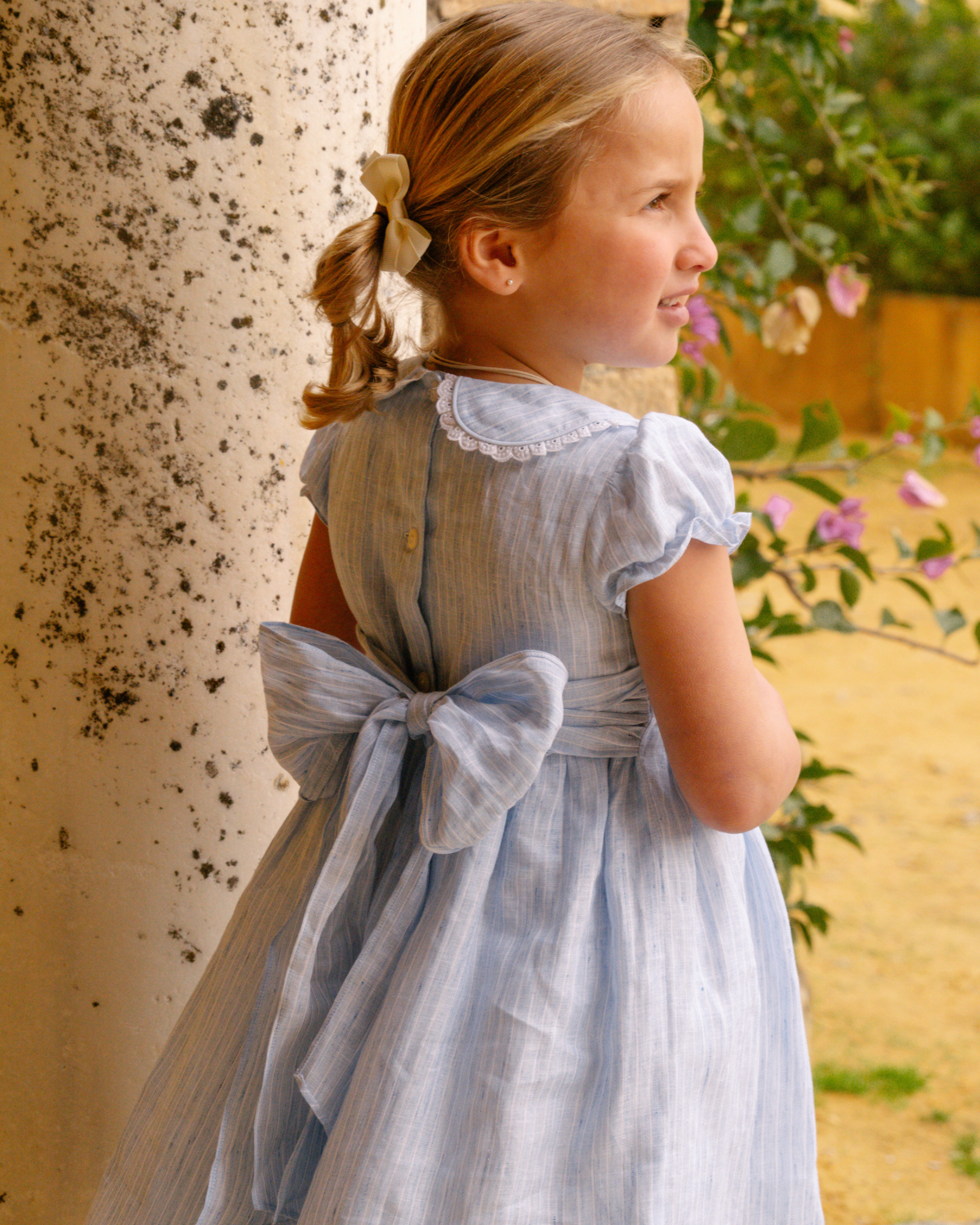 Young girl in a light blue dress with a bow standing outdoors.