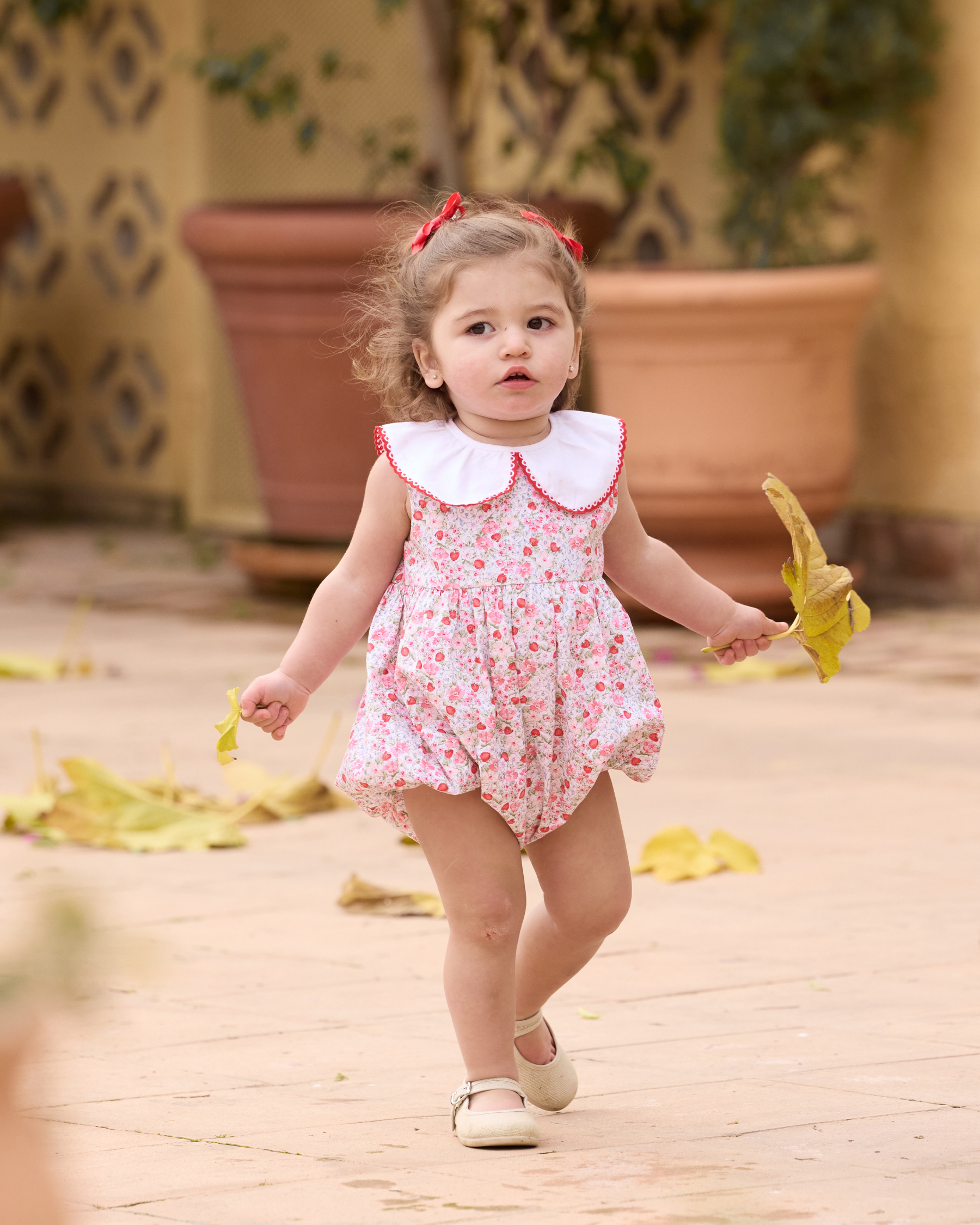 Young child in a floral dress walking outdoors with potted plants in the background