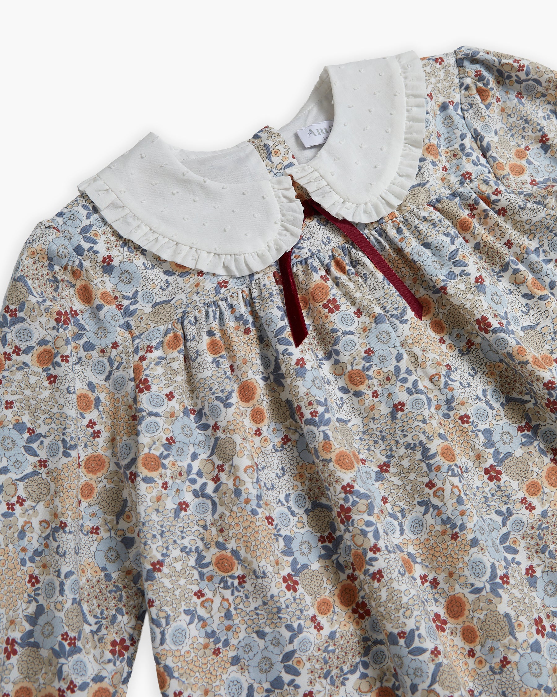 A close-up of a long-sleeve baby dress featuring a delicate floral and botanical print in muted blue, orange, and beige tones, with a contrasting white ruffled Peter Pan collar and a dark red tie at the neckline.
