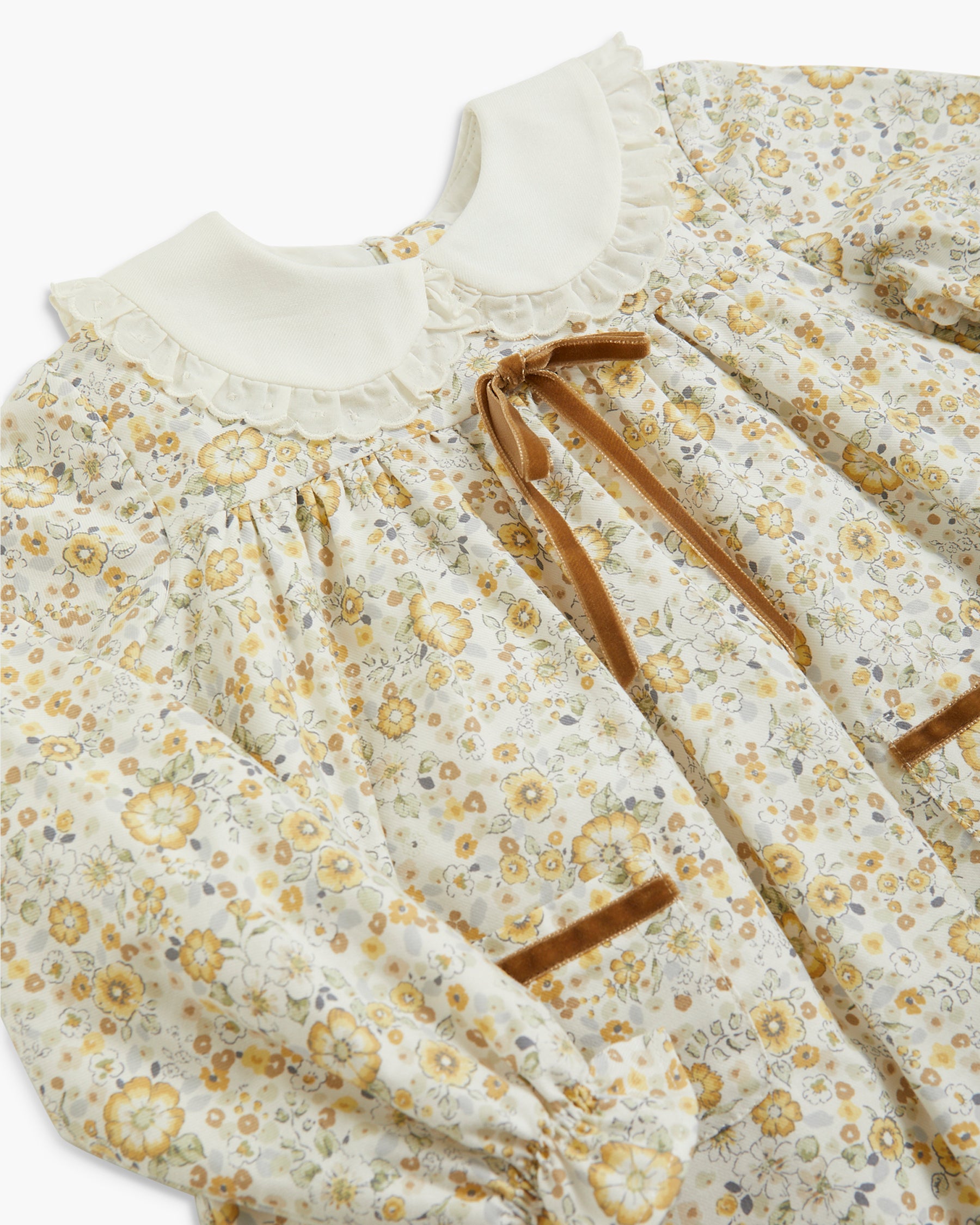 A close-up of a long-sleeve blouse featuring a cream-colored ruffled Peter Pan collar, a yellow and brown floral print, and brown ribbon ties at the chest.