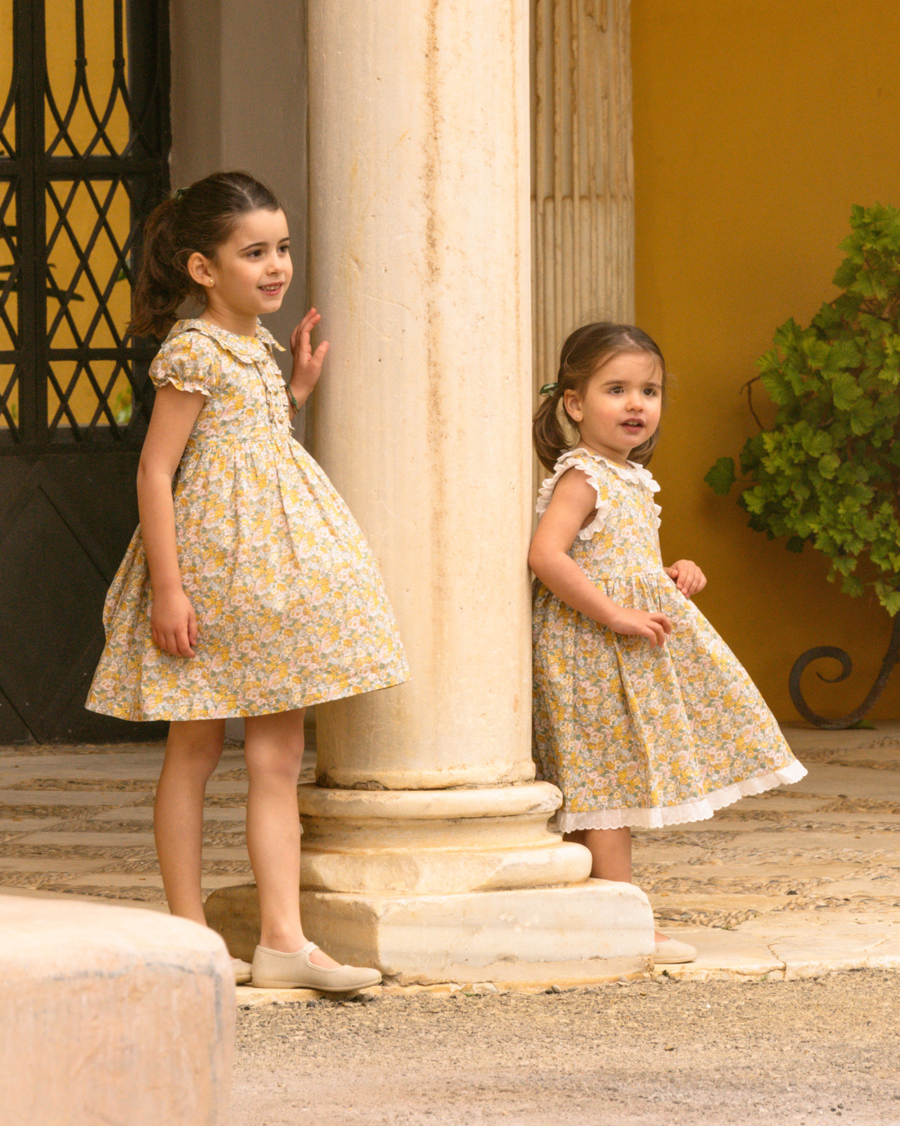 Two young girls in matching yellow floral dresses standing outdoors near columns.