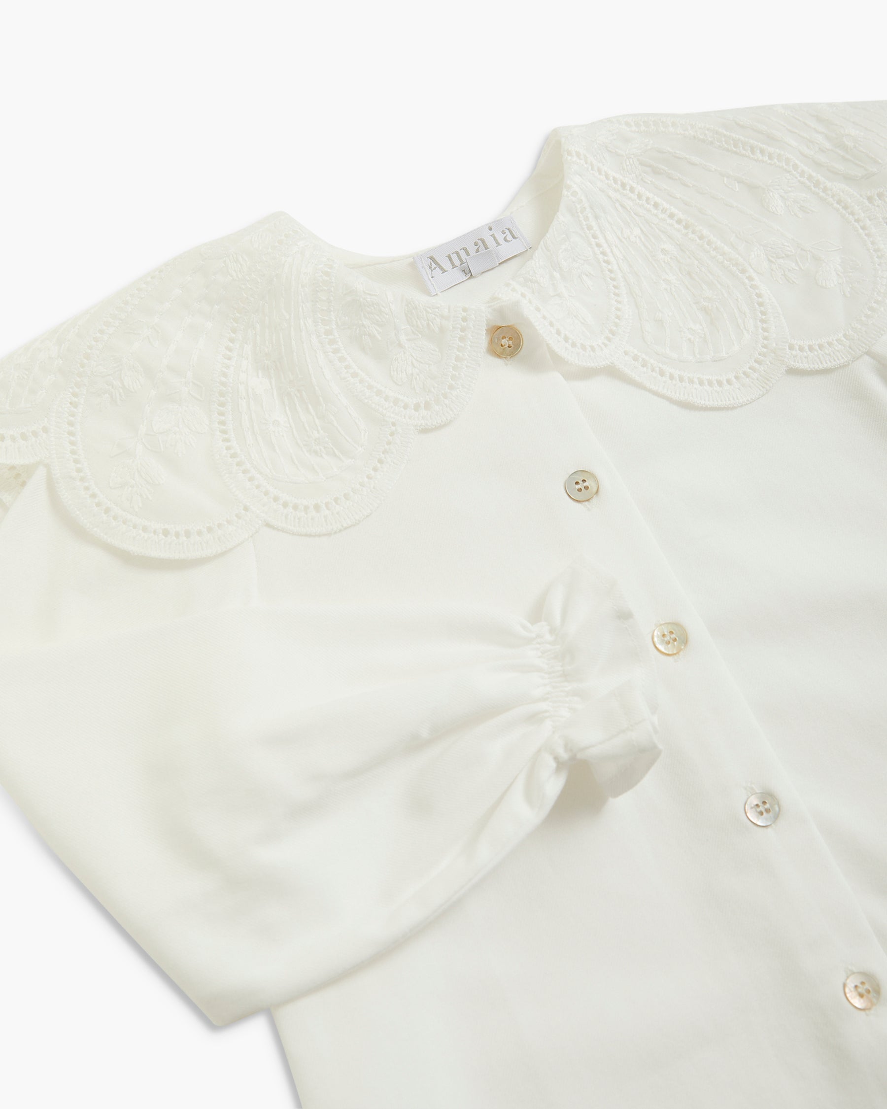 An off-white baby blouse featuring a large, decorative embroidered lace Peter Pan collar, a gathered front button placket, and a matching fabric bow on the chest. The brand name 'Amalia' is visible on the inner neck label.