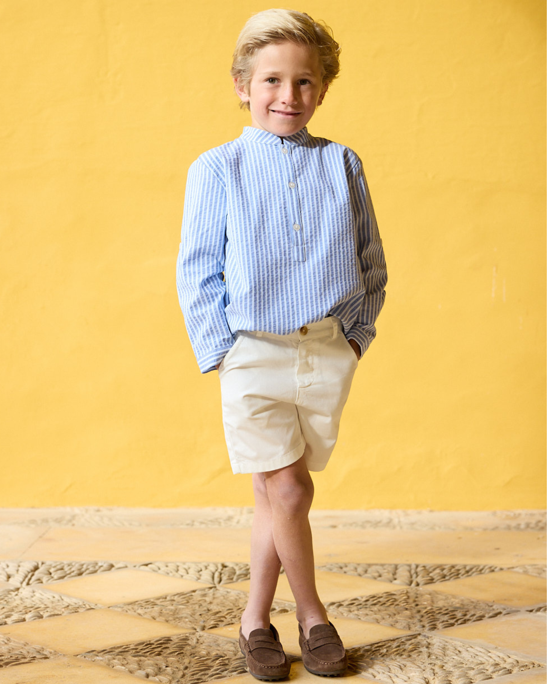 Young boy wearing a blue striped shirt and beige shorts against a yellow background