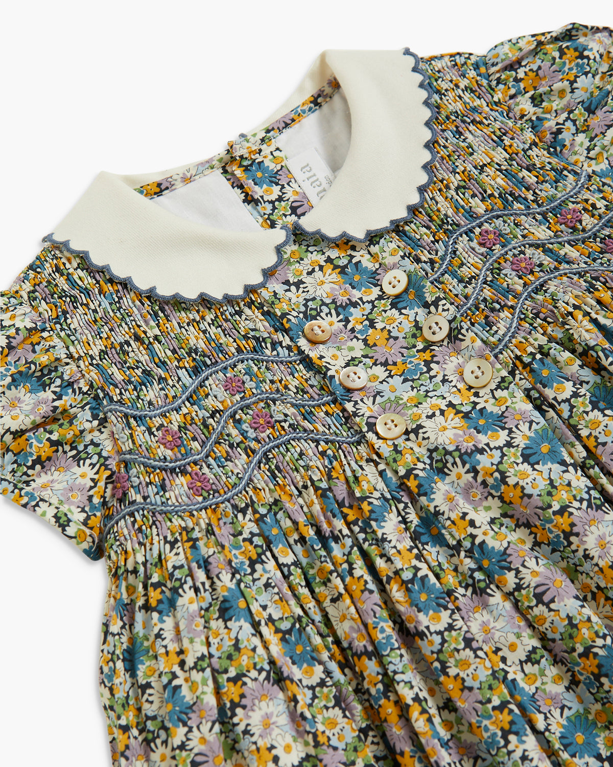 A close-up of a children's dress featuring a multicolor floral print, smocked bodice, scalloped cream collar, and decorative buttons.