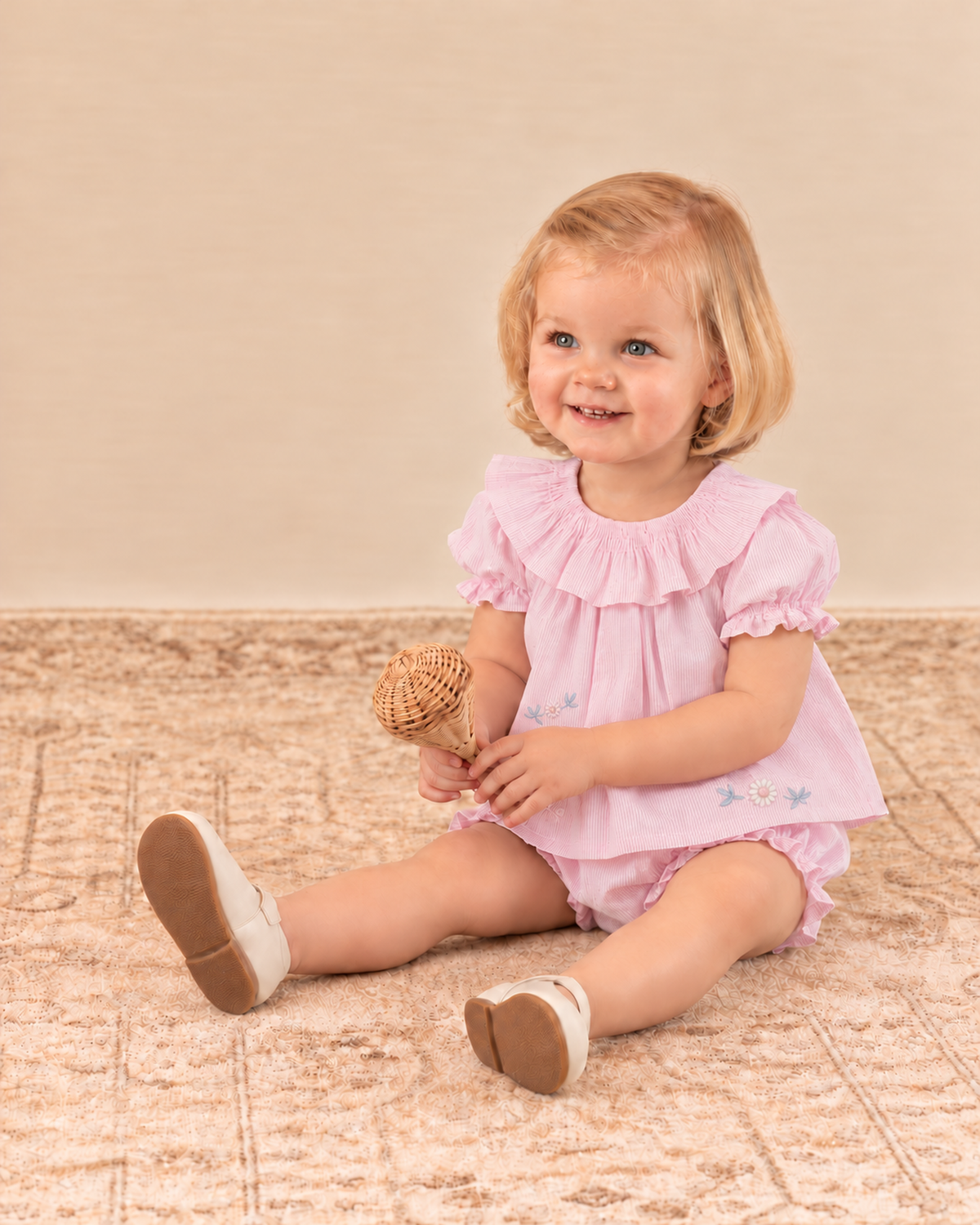 A young toddler girl with blonde hair sits on a patterned beige carpet, wearing a light pink ruffled short-sleeve romper dress and white shoes, holding a small woven toy.