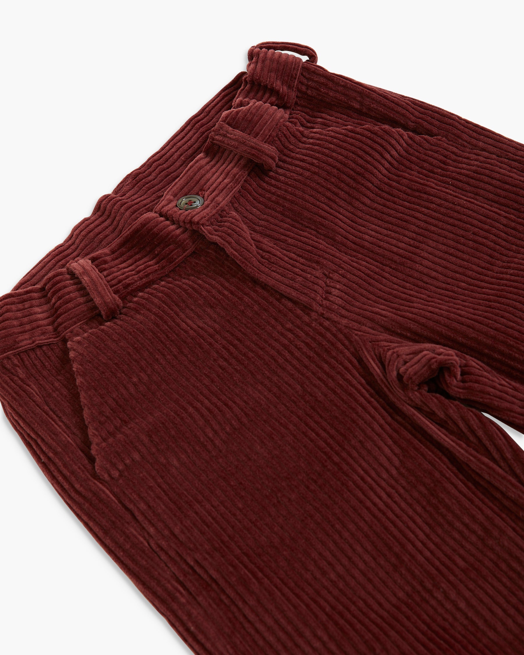 Close-up of dark burgundy corduroy trousers showing the ribbed texture, belt loops, and button closure on a white background.