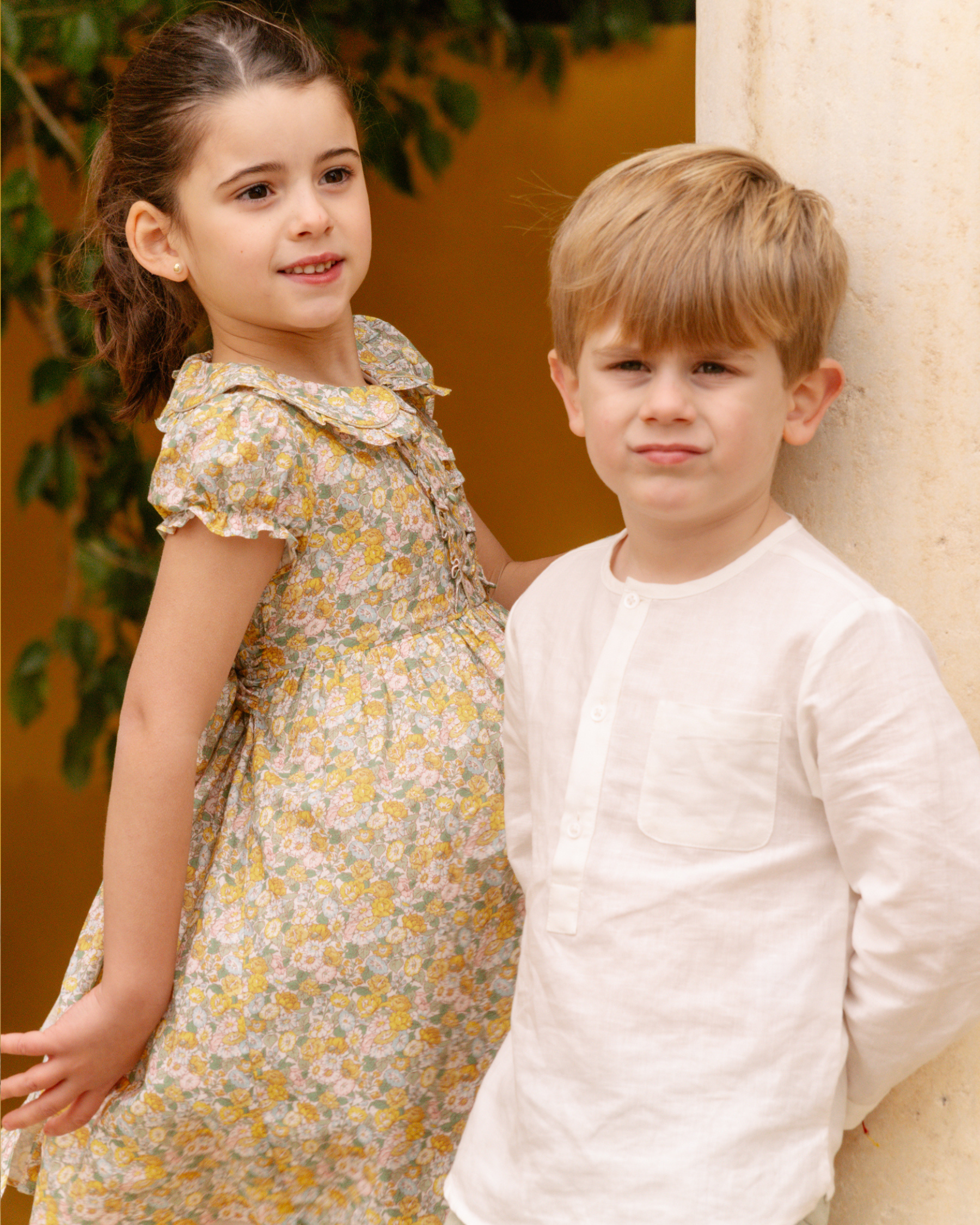 Two children, a girl in a floral dress and a boy in a white shirt, standing outdoors.
