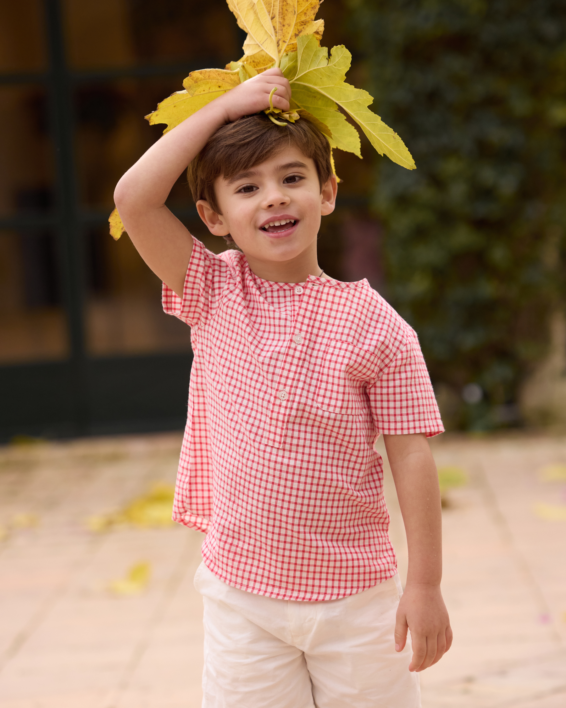 Child wearing a red checkered shirt and white pants, holding leaves above their head outdoors.