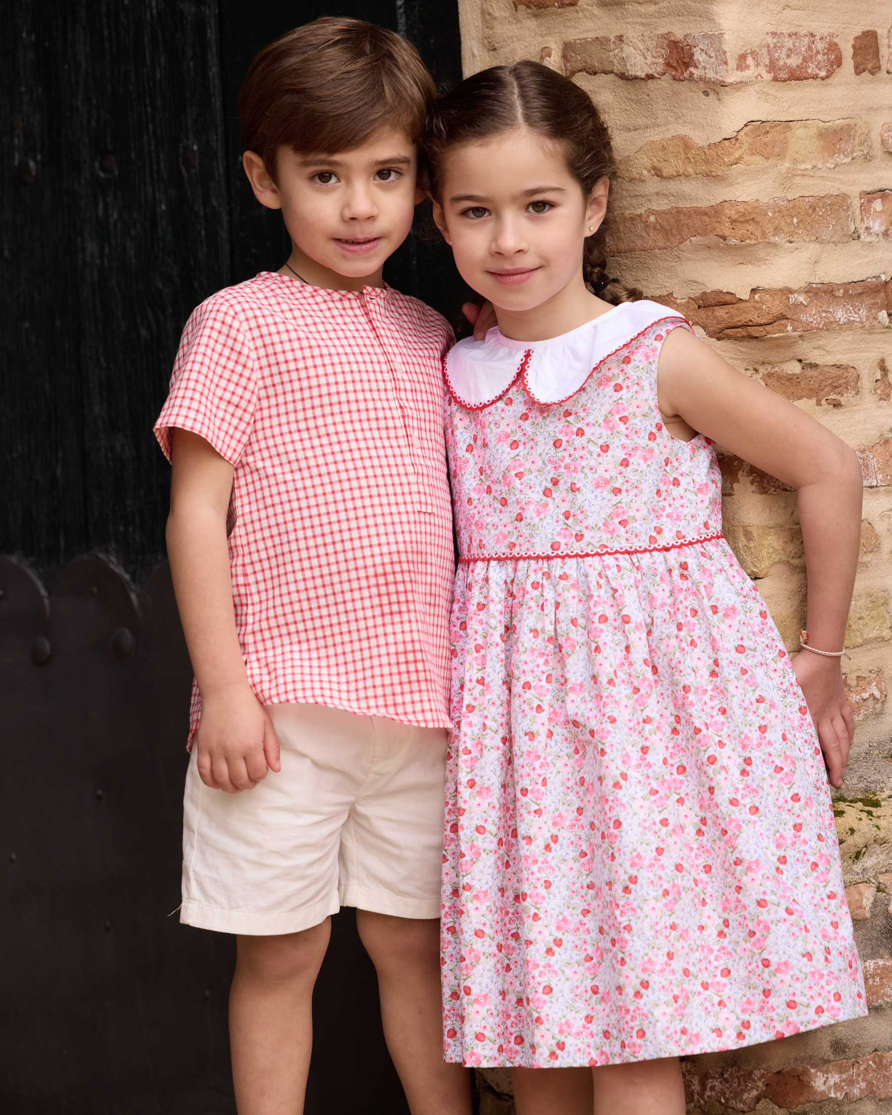 Two children in matching red and white outfits standing against a brick wall.